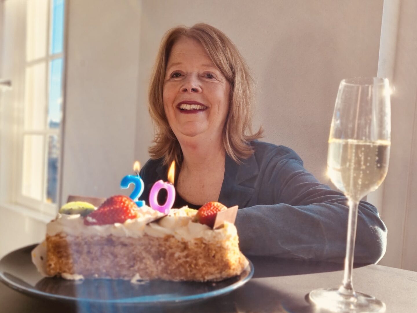 Olga zit aan tafel met een slagroomtaart voor zich. Daarom branden kaarsjes met het getal '20'. Naast haar staat een glas champagne. 